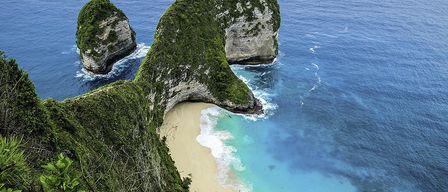 Ein Blick von oben auf zwei große Felsformationen mit Grün über einem sandigen Strand, umgeben von blauem Wasser.