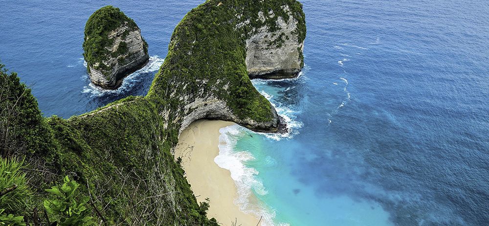 Ein Blick von oben auf zwei große Felsformationen mit Grün über einem sandigen Strand, umgeben von blauem Wasser.