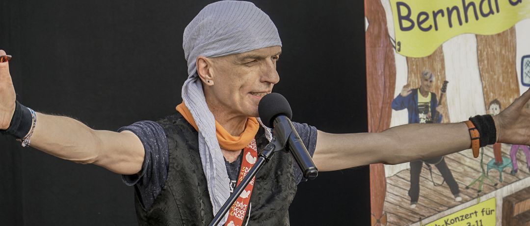 Ein Mann singt in ein Mikrofon und hält eine Gitarre, trägt ein graues Kopftuch und eine schwarze Weste. Hinter ihm ist ein Hintergrund mit einer Illustration von Kindern und einem gelben Banner mit weißem Text.