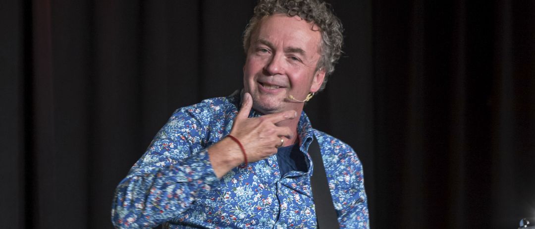 Ein Mann mit lockigem Haar tritt auf der Bühne auf. Er hält eine Gitarre mit der linken Hand und gestikuliert mit der rechten. Er trägt ein blaues Blumenhemd und Jeans.