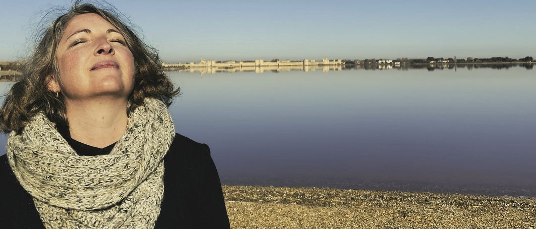 Eine Frau steht am Strand, den Himmel betrachtend, mit einem friedlichen Gesichtsausdruck. Ein großer Wasserkörper spiegelt entfernte Gebäude wider.