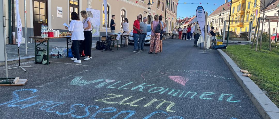 Eine Kreidezeichnung auf der Straße sagt 'Willkommen zum Dorffest'. Mehrere Leute stehen vor einem Gebäude. Einige Tische haben Gegenstände darauf.