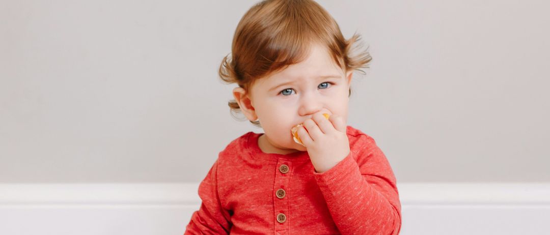 Ein Baby sitzt mit einem Obstteller auf dem Boden und isst eine Orangenscheibe. Es trägt ein rotes Hemd und graue Hosen.