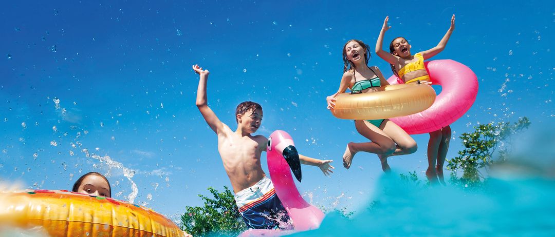 Vier Kinder spielen im Pool und tragen Schwimmwesten. Zwei Mädchen springen ins Wasser, während ein Junge einen rosa Schwimmkörper hält. Der Pool ist von Bäumen umgeben.