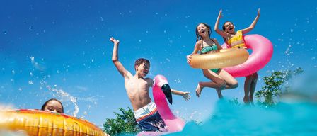 Vier Kinder spielen im Pool und tragen Schwimmwesten. Zwei Mädchen springen ins Wasser, während ein Junge einen rosa Schwimmkörper hält. Der Pool ist von Bäumen umgeben.
