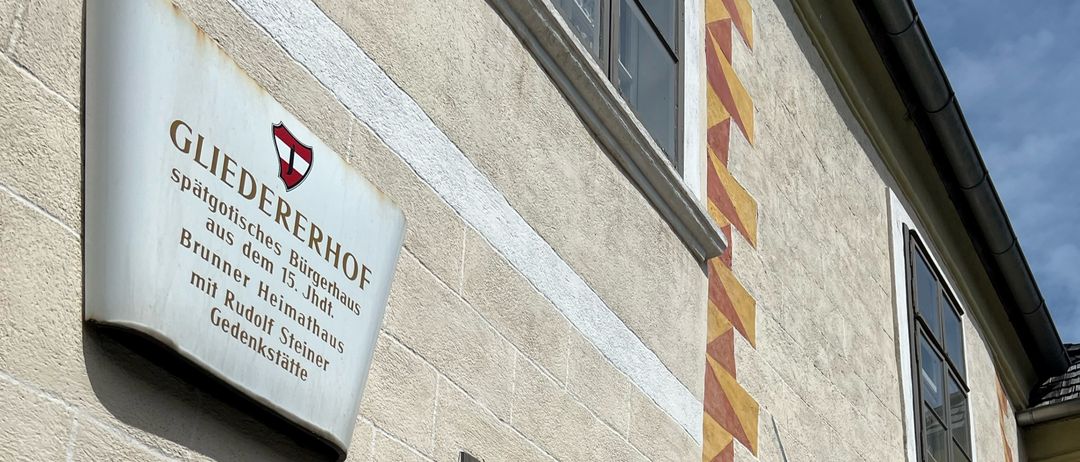 Ein Gebäudefassade mit einem Schild, das 'Gliedererhof spätgotisches Bürgerhaus auf dem 15. Jhd. Brunnter Heimathaus mit Rudolf Steiner Gedenkstätte' besagt. Unten am Schild befindet sich ein Fenster und die Tür ist braun mit einem Bogendesign.