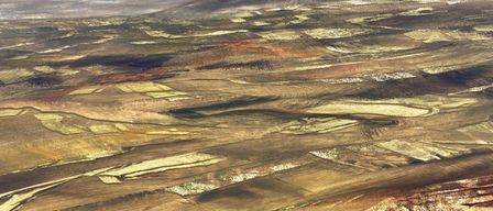 Luftaufnahme einer weiten, trockenen Landschaft mit Flecken von kultivierten Feldern und verstreuter Vegetation. Das Gelände zeigt wellige Hügel mit einer Mischung aus Braun-, Gold- und Grüntönen. Der Himmel ist bedeckt.