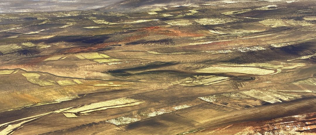 Luftaufnahme einer weiten, trockenen Landschaft mit Flecken von kultivierten Feldern und verstreuter Vegetation. Das Gelände zeigt wellige Hügel mit einer Mischung aus Braun-, Gold- und Grüntönen. Der Himmel ist bedeckt.