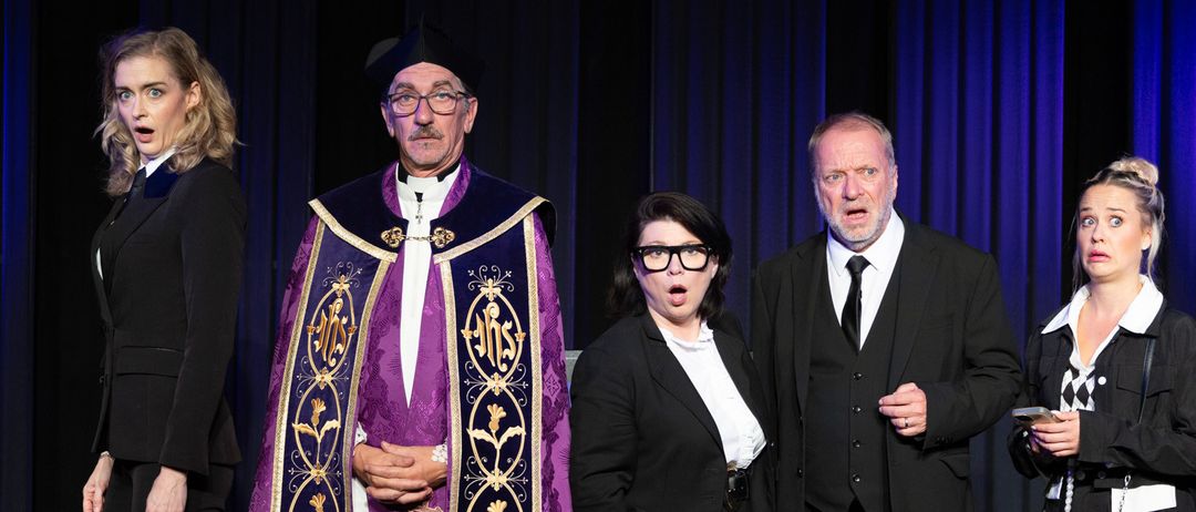 Fünf Personen in formeller Kleidung stehen auf einer Bühne. Ein Mann in einem lila-goldenen Gewand steht in der Mitte, während andere um ihn herum stehen. Der Hintergrund scheint eine Bühne mit blauen Vorhängen zu sein.