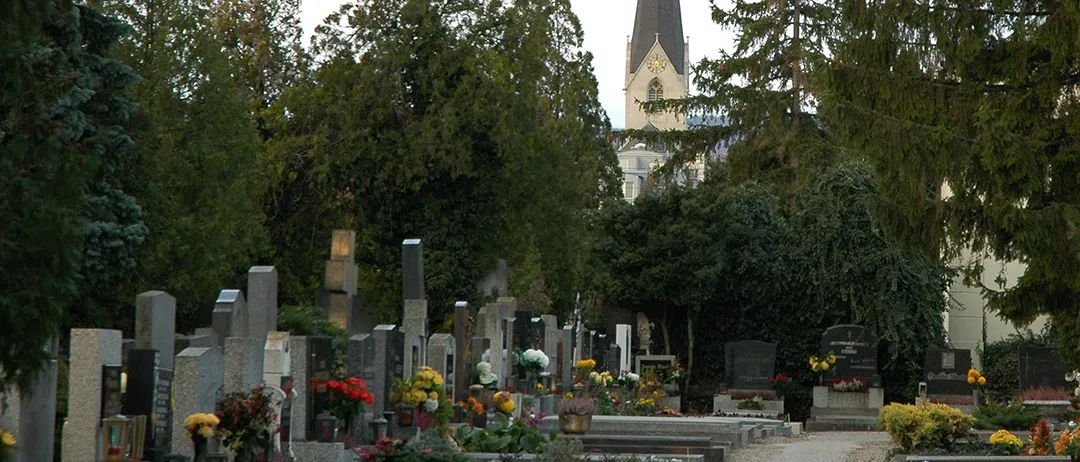 Ein Friedhof mit einem Kirchturm im Hintergrund. Der Friedhof ist mit Grabsteinen gefüllt und von Bäumen umgeben.