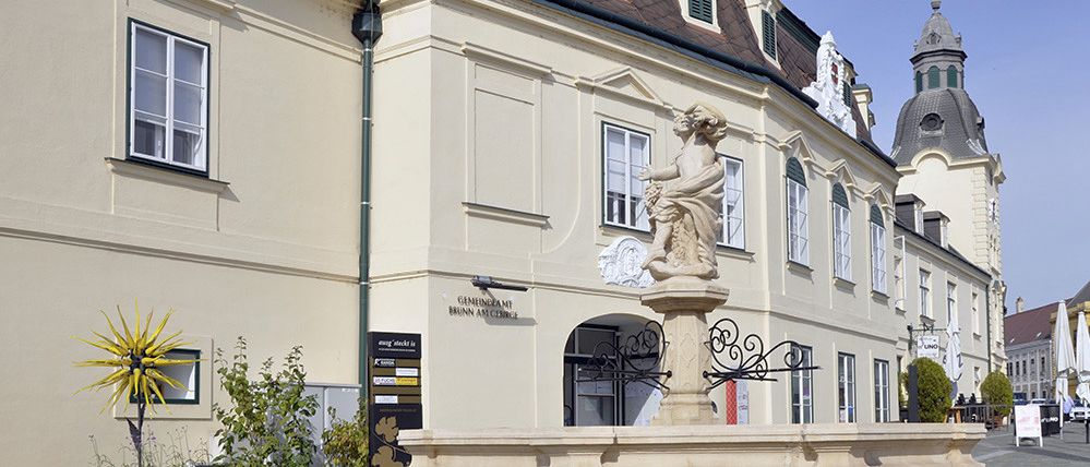 Ein beigefarbenes Gebäude mit mehreren Fenstern und einer Statue in der Mitte. Die Statue ist von einer Frau mit einem Kind auf ihren Schultern. Das Gebäude hat ein kuppelförmiges Dach und ist von Pflanzen umgeben.