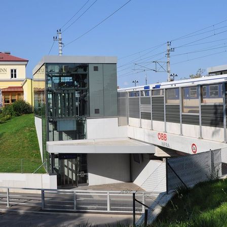Ein Bahnhof mit einem gläsernen Aufzug und einem vorbeifahrenden Zug. Der Bahnhof hat ein modernes Design mit einer weißen Überdachung und einem Zaun. Das Wort 'OBB' ist auf dem Bahnsteig geschrieben. Ein Gebäude mit rotem Dach befindet sich hinter dem Bahnhof.