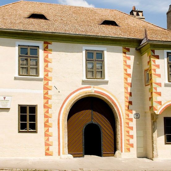 Ein altes Haus mit einem Ziegeldach und einem gewölbten Holztor. Das Haus hat mehrere Fenster und dekorative geometrische Muster an den Wänden. Über der Tür befindet sich die Nummer 34 an der Wand.
