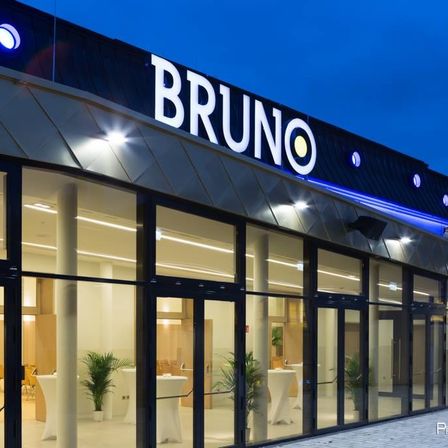 Das Äußere eines modernen Gebäudes mit dem Namen Bruno oben. Es hat Glaswände und -türen mit Pflanzen und Tischen im Inneren.