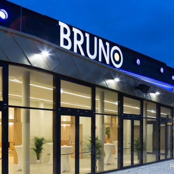 Das Äußere eines modernen Gebäudes mit dem Namen Bruno oben. Es hat Glaswände und -türen mit Pflanzen und Tischen im Inneren.