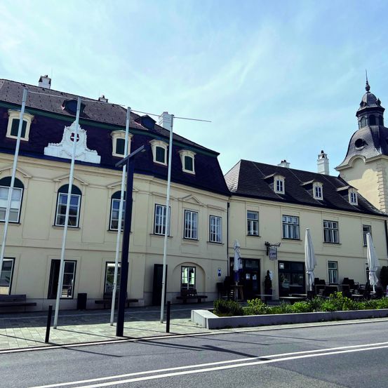 Ein großes beiges Gebäude mit schwarzen Dächern und mehreren Fenstern. Es hat eine Flagge oben und eine Außensitzgelegenheit mit Schirmen.