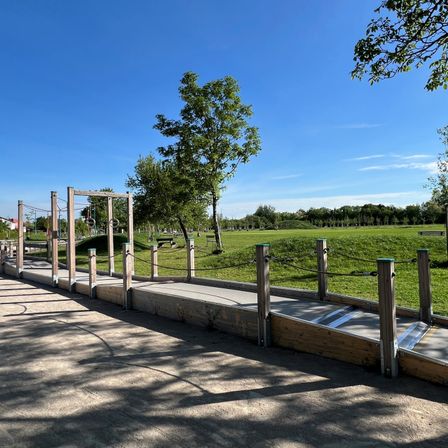 Ein Spielplatz mit Schaukeln und einer Brücke, umgeben von Bäumen, unter einem klaren blauen Himmel.