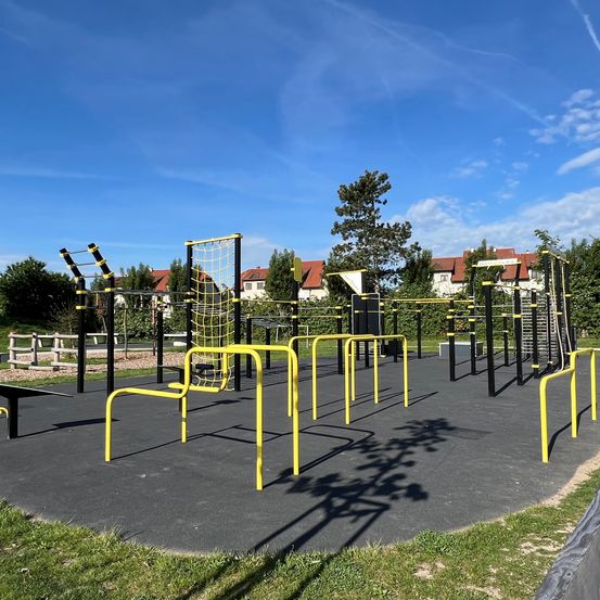 Ein Freiluft-Fitnessstudio mit gelben und schwarzen Geräten. Bäume und Häuser mit roten Dächern im Hintergrund.