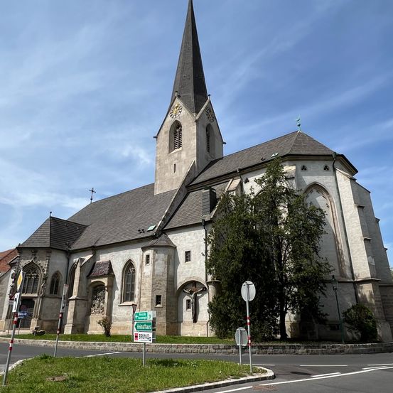 Eine alte Kirche mit Turm und Uhr steht neben einer Straße, mit einem großen Baum davor.
