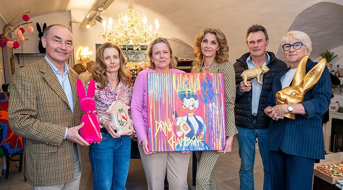 Fünf Personen posieren für ein Foto in einem Raum mit einem Kronleuchter. Sie halten Geschenke, darunter ein rosa Hase, ein goldener Hase und ein Gemälde.