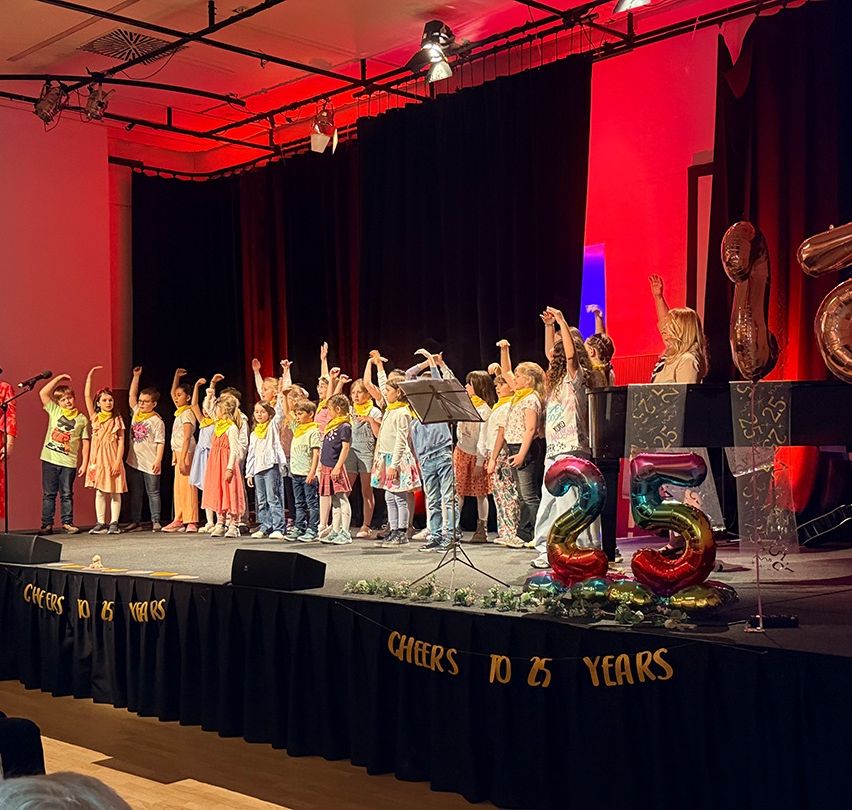 Eine Gruppe von Kindern in gelben Schals tritt mit einem 'Cheers to 25 Years'-Banner auf der Bühne auf, begleitet von einer Frau und Luftballons.