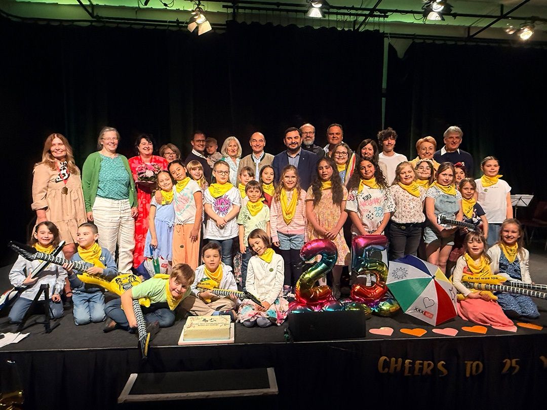 Eine Gruppe von Menschen auf der Bühne, die mit Luftballons und einem Kuchen feiern. Einige halten Gitarren, und es gibt ein Schild, das Cheers To 25 sagt.