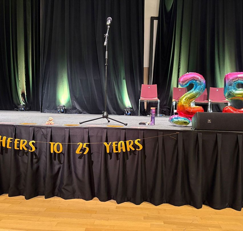 Eine Bühne mit einem Mikrofon auf einem Ständer, einem Banner mit der Aufschrift 'Jahre bis 25 Jahre' und Luftballons mit der Nummer 25 auf beiden Seiten. Schwarze Vorhänge sind zugezogen, und es gibt Stühle hinter der Bühne.