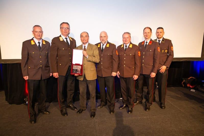 Sieben Männer in Uniformen stehen auf einer Bühne, einer hält einen Preis. Der Mann in der Mitte trägt eine Brille und einen braunen Mantel. Ein weißer Hintergrund ist hinter ihnen.