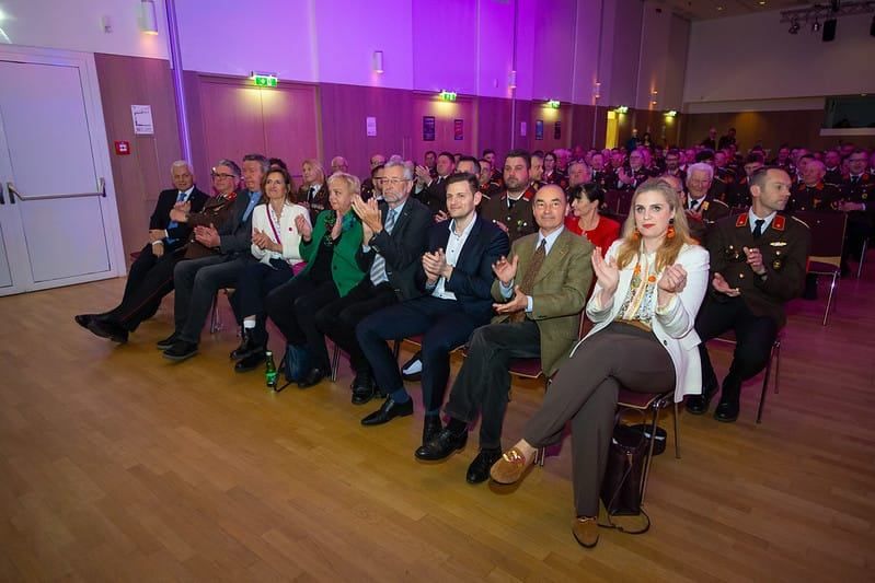 Ein großes Publikum sitzt in einem Saal und klatscht. Einige Personen tragen formelle Kleidung und Krawatten.