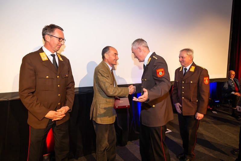 Vier Männer in Uniformen stehen vor einer Leinwand; zwei schütteln Hände, einer hält eine rote Schachtel.