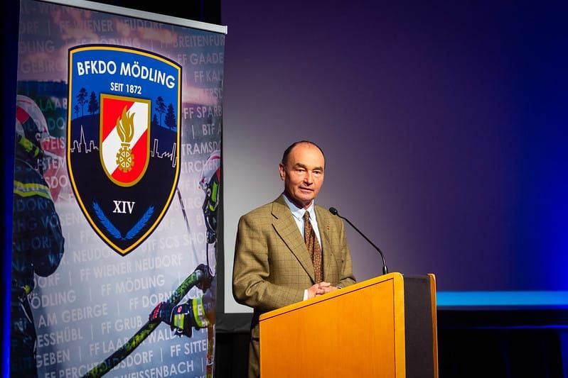 Ein Mann steht lächelnd am Podium mit einem Mikrofon. Dahinter zeigt ein großes Banner das Logo einer Feuerwehr, was auf eine Veranstaltung oder Ankündigung hinweist.