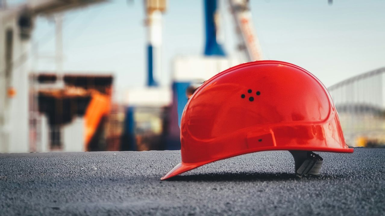 Ein roter Schutzhelm mit einigen Löchern auf der Oberseite liegt auf einer grauen Oberfläche in einem verschwommenen Hintergrund mit einem Kran.