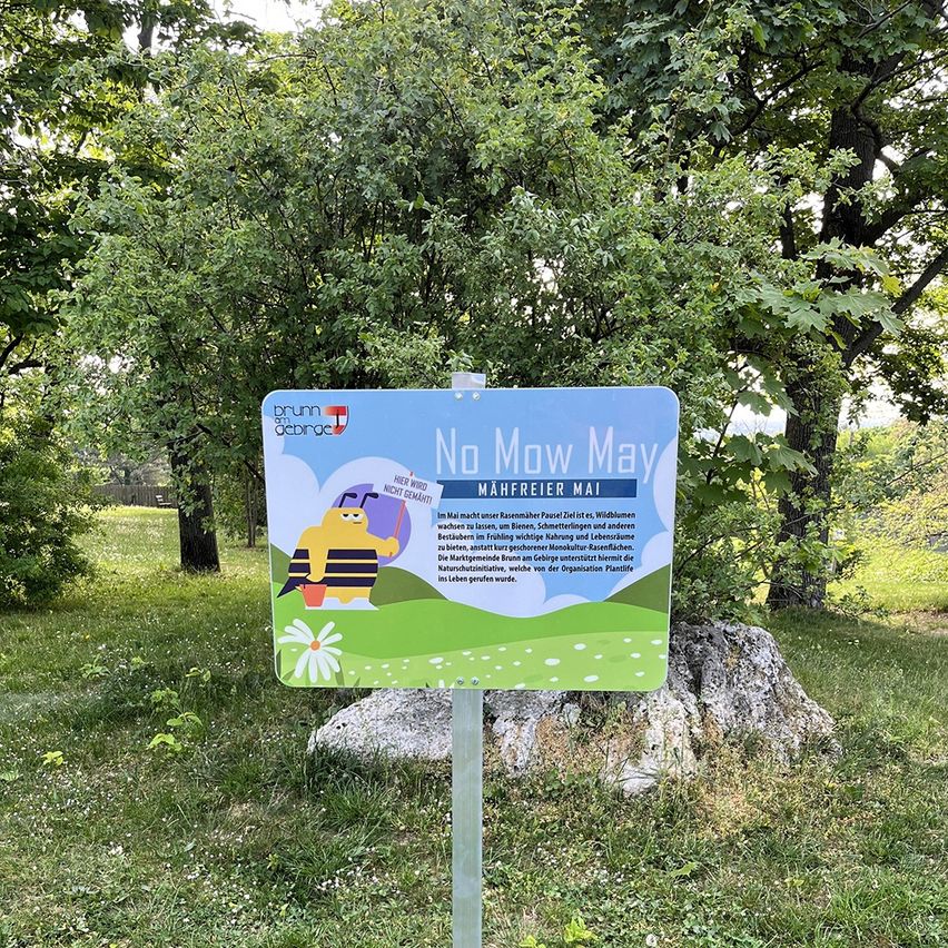 Ein Schild mit einer Biene-Figur steht 'Mähreier Mai' und ermutigt dazu, im Mai nicht zu mähen. Das Schild steht auf einem Rasen mit Bäumen.