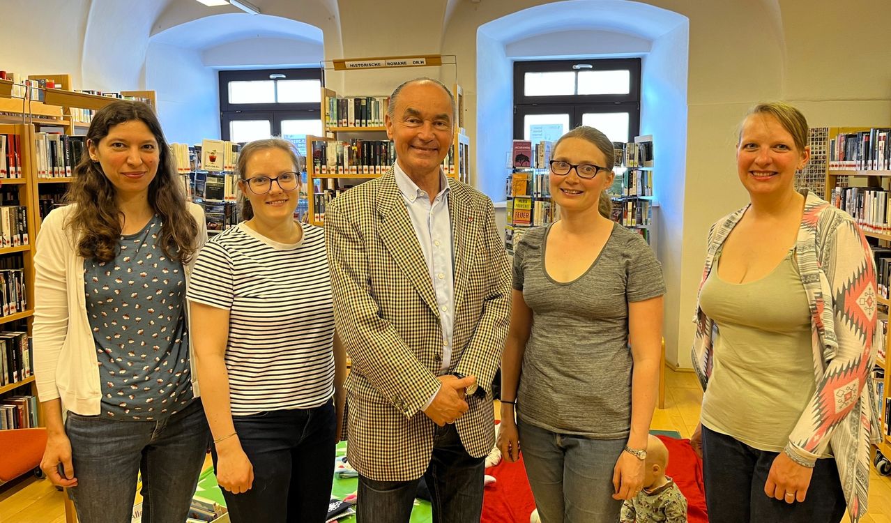 Vier Personen stehen in einer Bibliothek. Ein Mann in einem karierten Anzug steht zwischen zwei Frauen. Ein Baby liegt auf dem Boden.