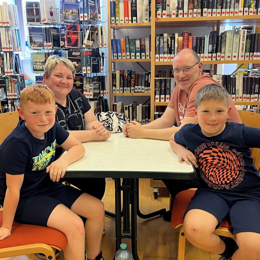 Eine vierköpfige Familie posiert für ein Foto an einem Tisch in der Bibliothek. Zwei Jungen sitzen einander gegenüber, während ihre Eltern lächelnd hinter ihnen stehen. Hinter ihnen sind mit Büchern gefüllte Regale zu sehen.