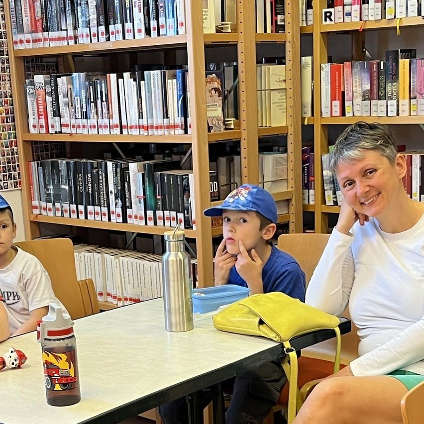 Eine Frau und zwei Kinder sitzen in einer Bibliothek an einem Tisch, umgeben von Regalen voller Bücher. Die Frau lächelt und ein Kind sieht nachdenklich aus.