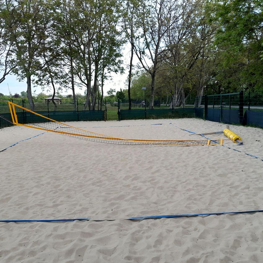 Ein Freiluft-Volleyballfeld mit gelben Netzen und blauen Begrenzungslinien auf einem sandigen Feld, umgeben von grünen Bäumen und Zäunen.