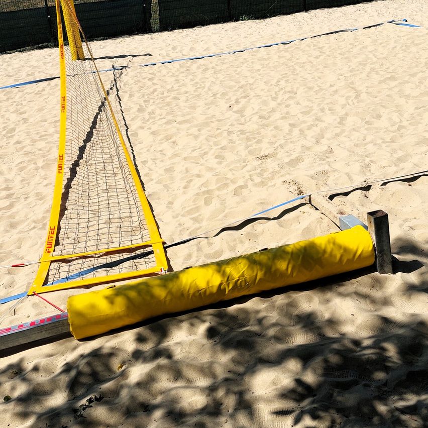 Ein gelbes Volleyballnetz steht auf einem Sandstrand, gesichert durch blaue Seile. Eine gelbe gepolsterte Barriere ruht auf dem Sand neben dem Netz.