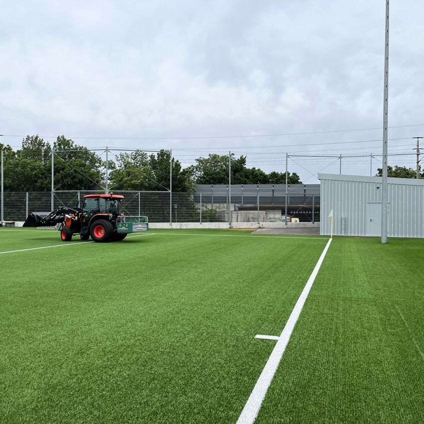 Ein leeres Sportfeld mit grünem Rasen. Ein roter Traktor ist an der Seite geparkt, und eine weiße Linie ist auf dem Feld gezogen. Ein Zaun umgibt das Feld.
