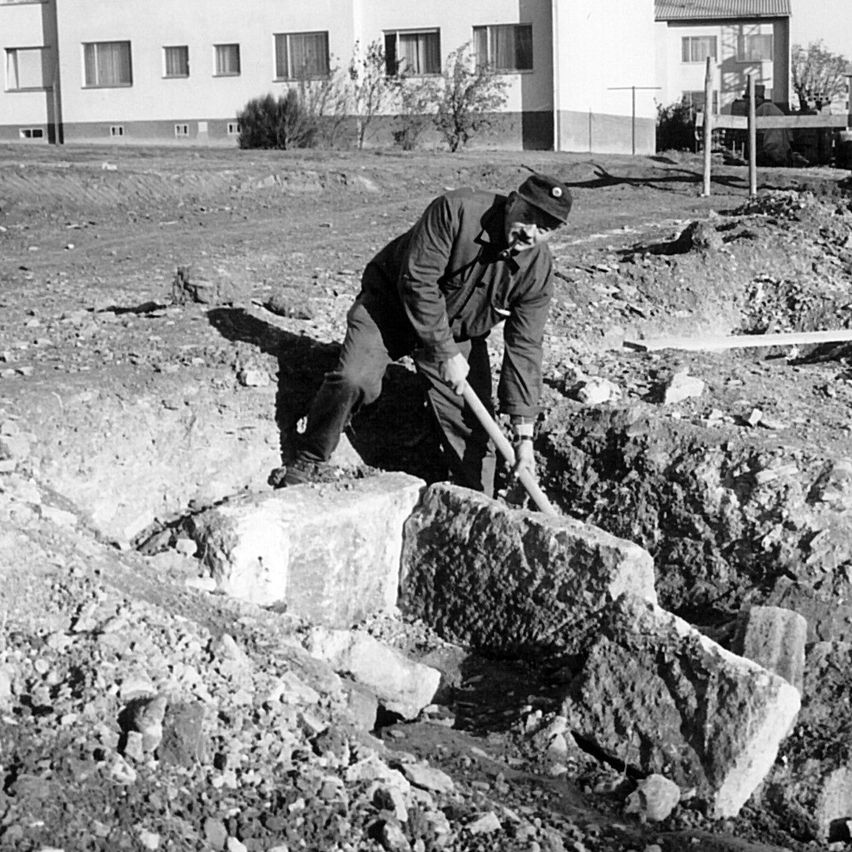 A black and white image of a man digging a hole with a shovel in a dirt field with a building in the background.