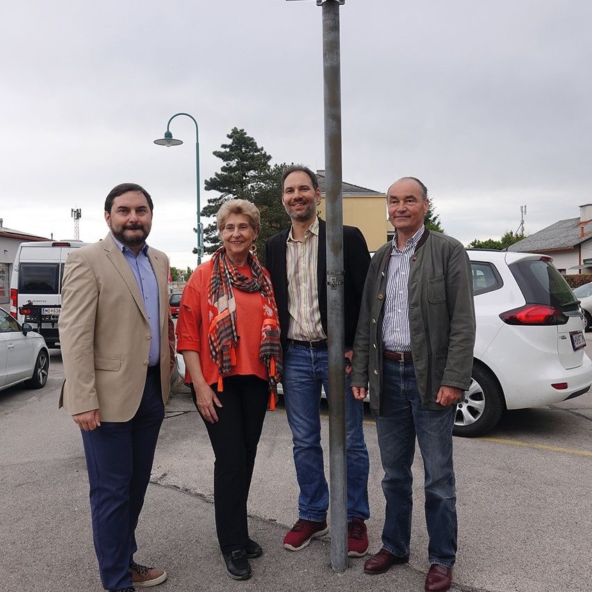 Four people stand in a parking lot near a pole. A man in a beige coat and blue pants is on the left.