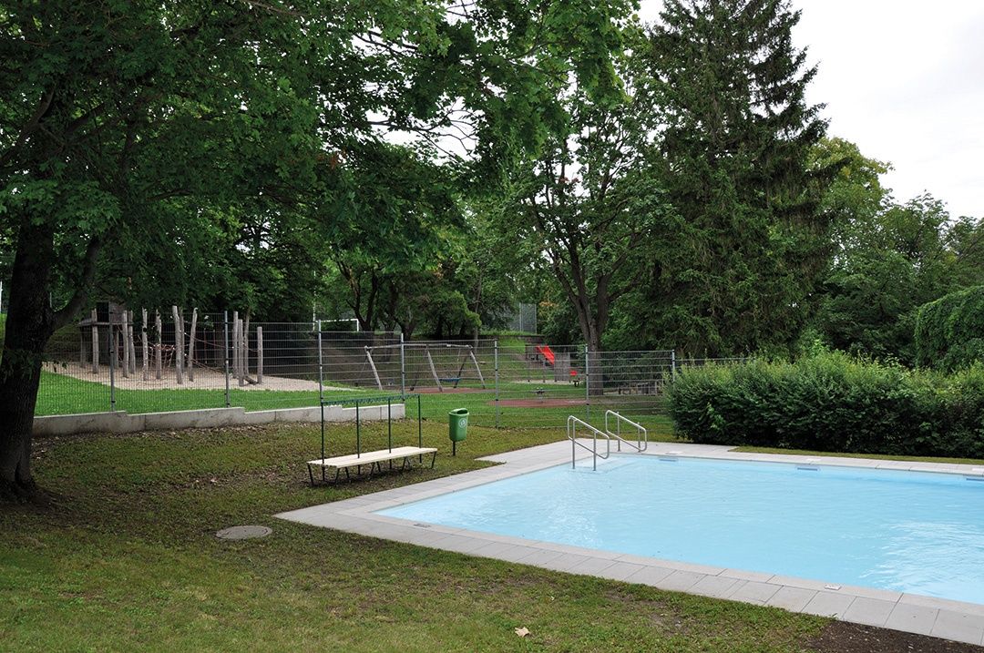 An empty outdoor pool with a bench and a trash can, surrounded by a grassy area and a fence with a playground.