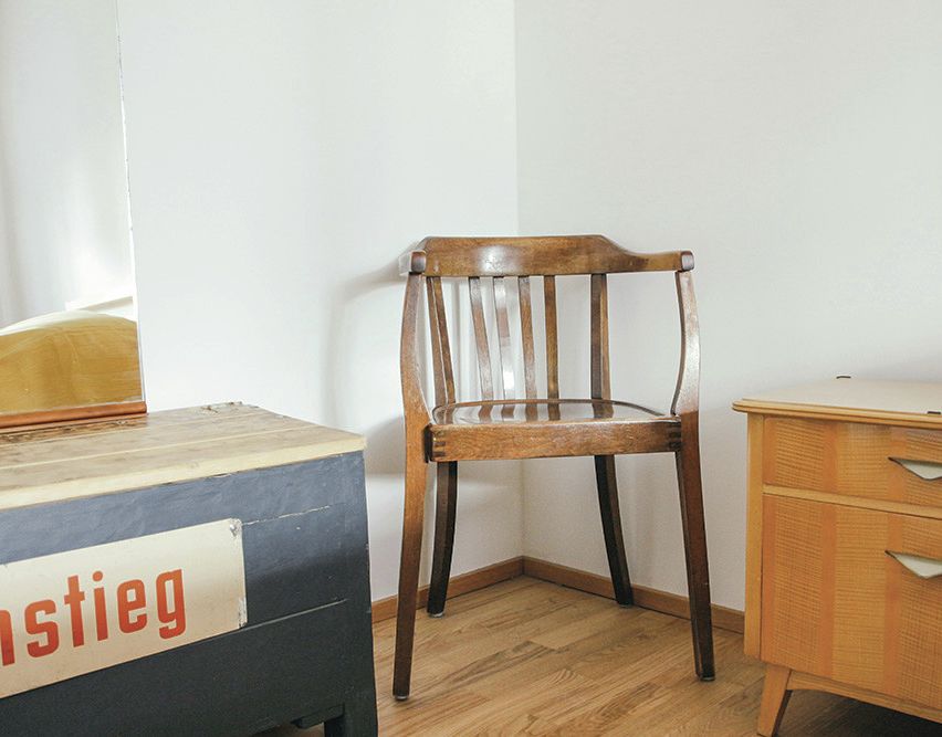 Ein Holzstuhl steht in einer Ecke eines Zimmers mit weißen Wänden. Ein Holzschreibtisch mit Schubladen befindet sich rechts, und eine Aufbewahrungsbox mit einem roten Etikett, das 'tieg' liest, steht links. Ein Spiegel ist an der linken Wand.