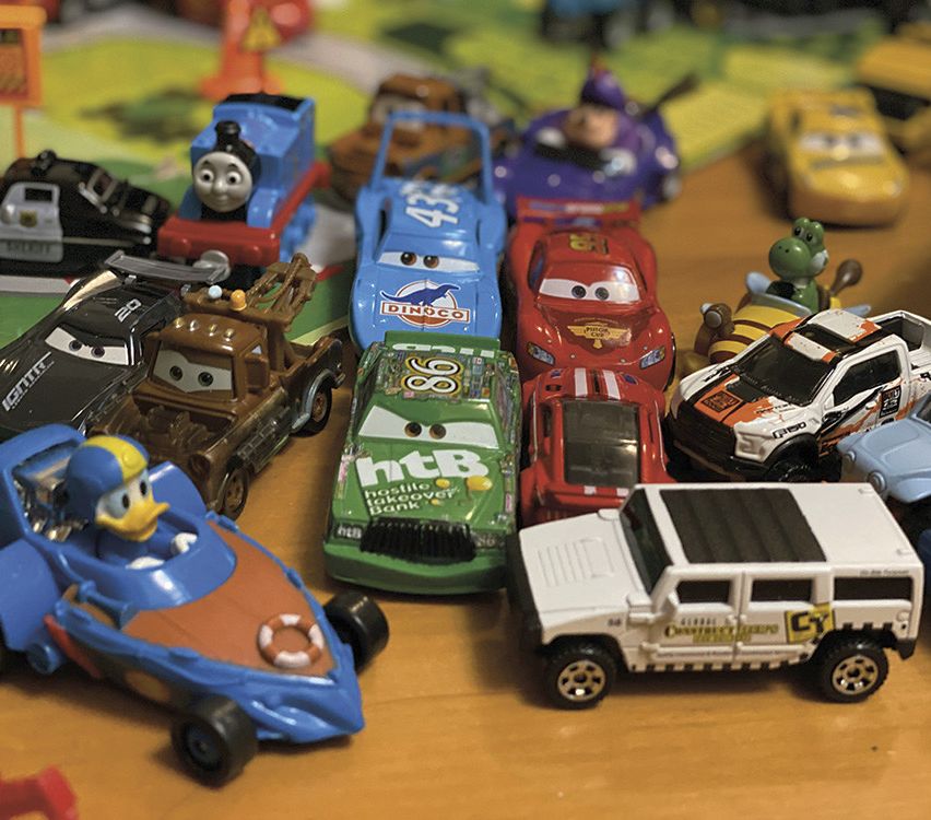 Eine Sammlung von Spielzeugautos aus verschiedenen Serien, einschließlich eines blauen Bootes und eines roten Autos, befinden sich auf einer braunen Oberfläche. Sie umfassen Disneys Cars mit Charakteren wie Lightning McQueen, Mater und Sally.