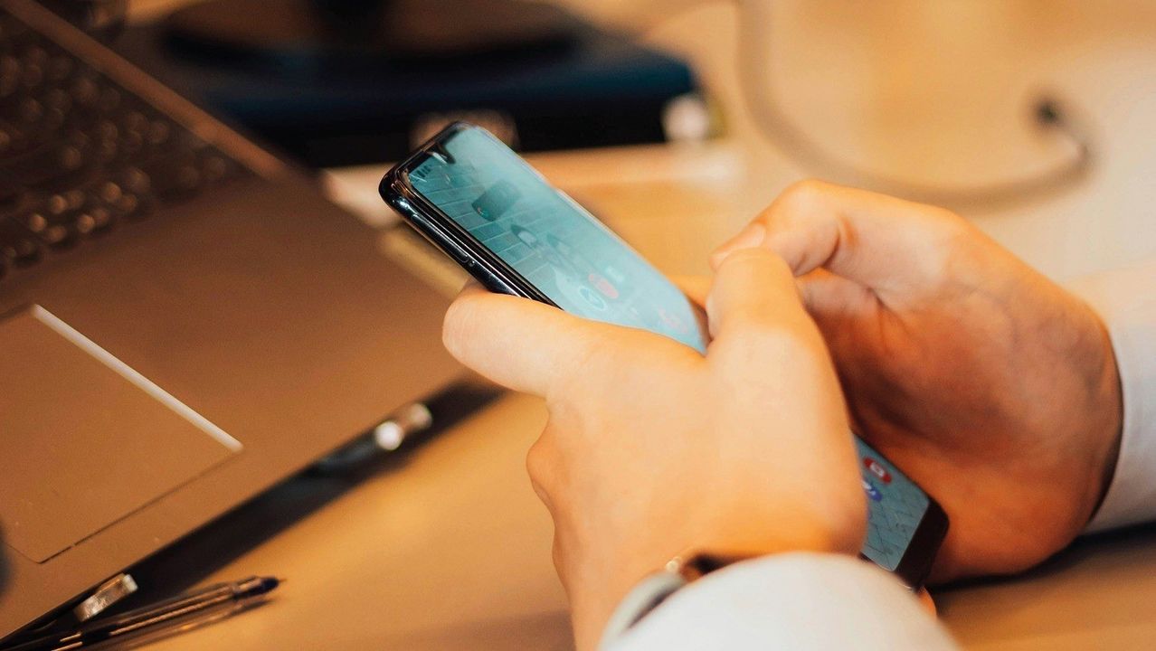 Eine Hand hält ein Smartphone, das ein Bild eines Gebäudes und eine Karte zeigt. Der Hintergrund zeigt einen Schreibtisch mit einem Laptop und einem Stift.