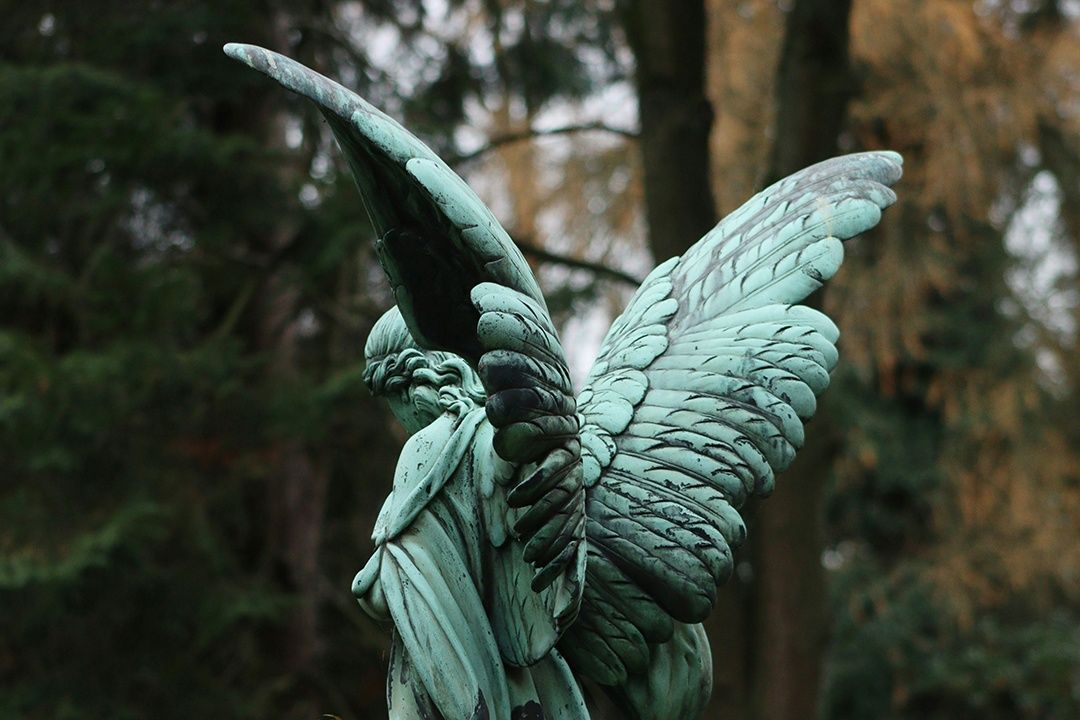 Engelstatue aus patiniertem Kupfer mit großen Flügeln vor Bäumen auf einem Friedhof