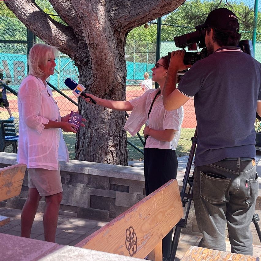 Eine Frau in einem rosa Hemd wird von einem Reporter interviewt, der ein Mikrofon hält. Der Reporter filmt mit einer Kamera. Hinter ihnen ist ein Baumstamm zu sehen, und im Hintergrund ist teilweise ein Tennisplatz zu erkennen.