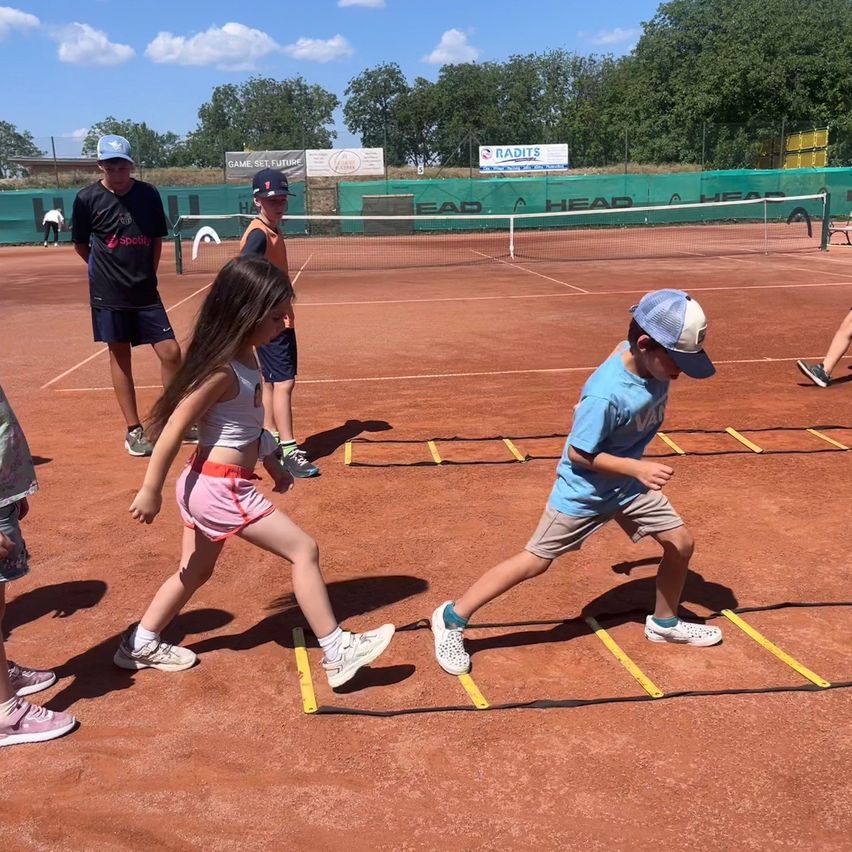 Kinder üben auf einem Tennisplatz Beweglichkeitsübungen, wobei eines der Kinder einen Hut und das andere eine Kappe trägt. Ein Tennisnetz ist im Hintergrund zu sehen.