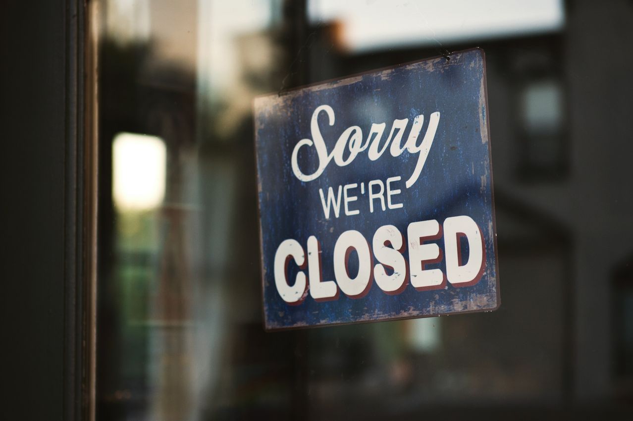 Ein blaues Schild mit weißen Buchstaben steht auf einer Glasscheibe und zeigt 'Entschuldigung, wir sind geschlossen'. Das Schild hat einen rustikalen Look mit abgenutzten Kanten.