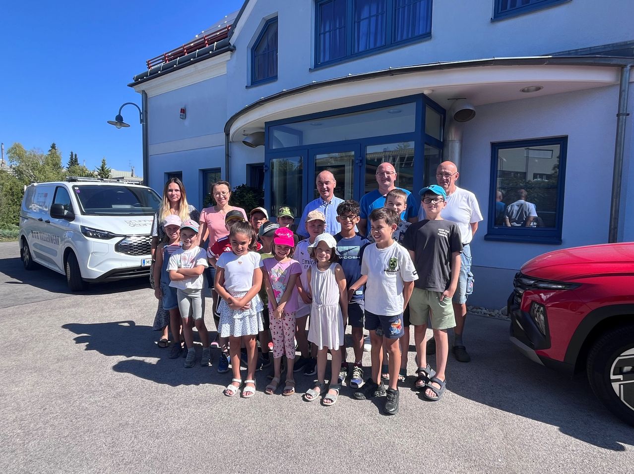 Eine Gruppe von Kindern und Erwachsenen steht vor einem Gebäude mit blauen Fenstern und einem roten Auto in der Nähe. Die Kinder tragen Hüte und Sandalen, während die Erwachsenen Brillen und Turnschuhe tragen.